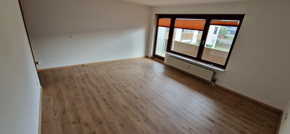 Leerer Raum mit Holzboden und großen Fenstern mit orangenem Sichtschutz.