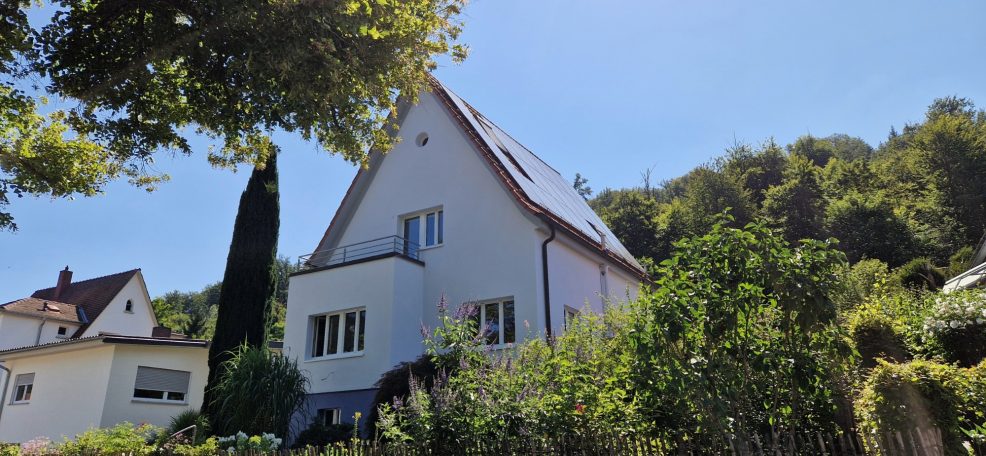 Helles Haus mit Garten, umgeben von Bäumen und Hügeln unter blauem Himmel.