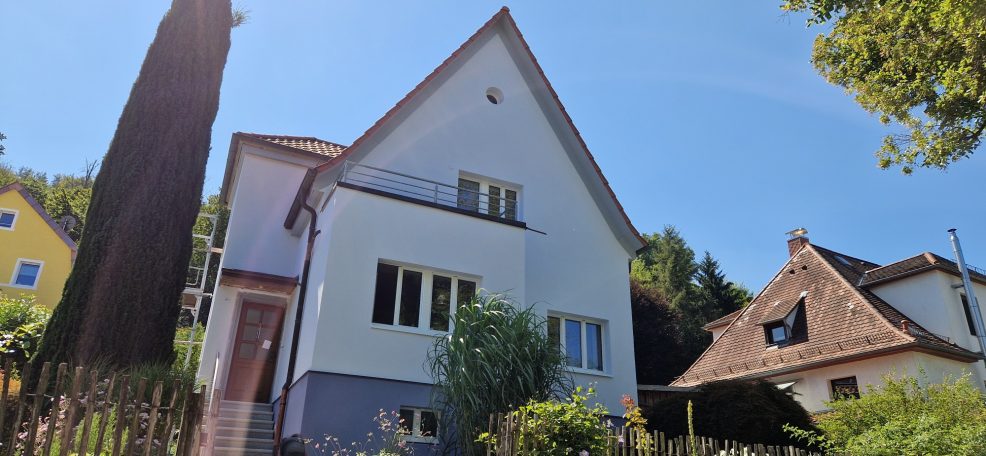 Zwei-geschossiges Wohnhaus mit Giebel, umgeben von Pflanzen und Bäumen.