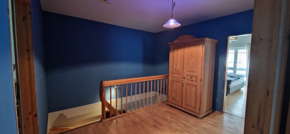Ein blauer Raum mit Holztreppe und Kleiderschrank, Zugang zu einem weiteren Raum.