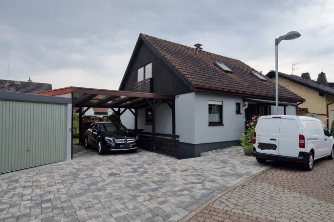 Ein Einfamilienhaus mit Carport und Fahrzeugen auf einem gepflasterten Grundstück.